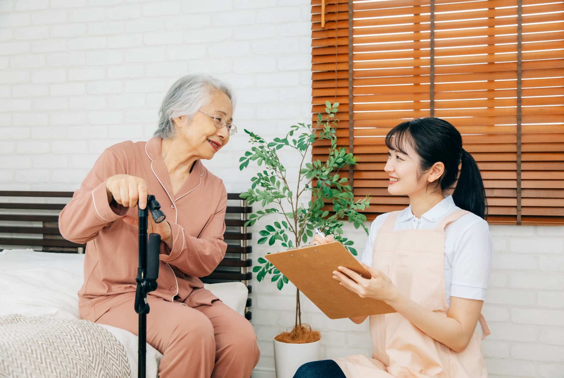 杖を持ってベッドに腰掛ける高齢女性と、その横にバインダーを持って語りかける女性介護士